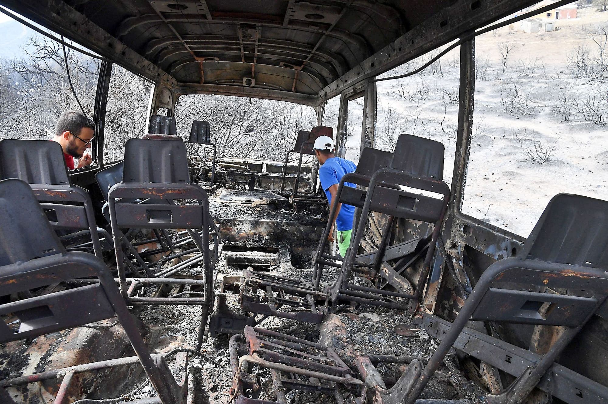 Deux hommes fouillent la carcasse complètement calcinée d’un autobus dans lequel 12 personnes auraient perdu la vie, piégées par un feu de forêt.